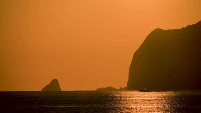 A silhouette of a fishing boat sailing past the volcanic Keelung Islet in Taiwan during a golden morning sunrise with orange sky and glowing sea.