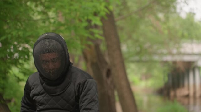Close up of person in black fencing gear standing outdoors near trees engaging in swordplay with another fencer. person is seen preparing and striking during activity wearing protective mesh