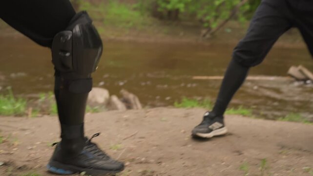 Close up of two people in black protective gear engaged in sword fight near water. One person wields long sword while other is partially visible with arm and leg armor. scene features