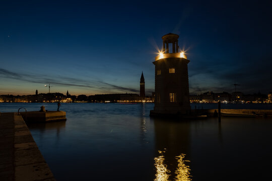 A lit stone lighthouse glows in the dark lagoon waters against the distant Venice skyline at twilight. Peaceful nocturnal views inspire romantic European travel and celebrate rich maritime heritage.