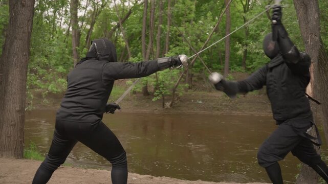 Two people in black fencing gear engage with swords near riverbank surrounded by trees. Their movements are fluid as they parry and thrust demonstrating skill against each other. natural