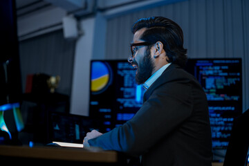 Fototapeta premium South Asian male developer in a dark jacket reads data on a tablet beside monitors streaming code in a blue-tinted tech office.