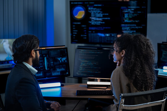 South Asian male and Black female cybersecurity analysts point at threat monitors discussing solutions in a dark operations center.