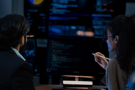 South Asian male and Black female software developers pair-program at dual monitors displaying network security algorithms at night.