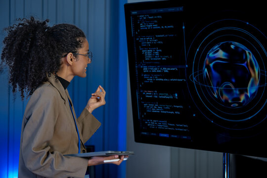 Black female cybersecurity analyst with curly hair and glasses types at a keyboard while code and data fill large screens behind her.
