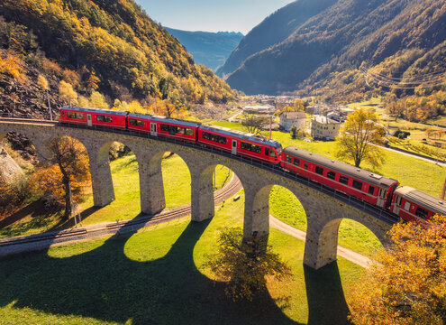 Aerial drone view of the red Bernina Express train crossing the Brusio Viaduct in Switzerland on sunny autumn day, colorful trees, alpine village in mountain valley scenery. Top view of train in Alps
