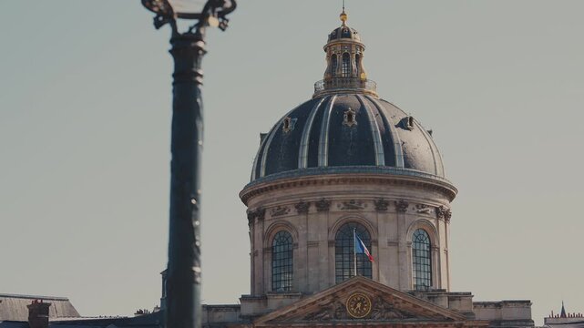 Smooth tilt up shot revealing the grand dome of the Institut de France with the French national flag waving, shot from Pont des Arts in Paris, France. High quality 4K B-roll video.