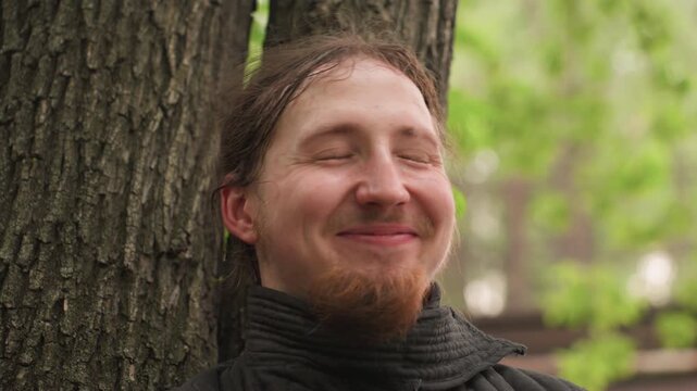 Close up of man with light beard and dark hair wearing black high collar jacket leaning against tree trunk in outdoor park setting soft natural lighting green foliage background