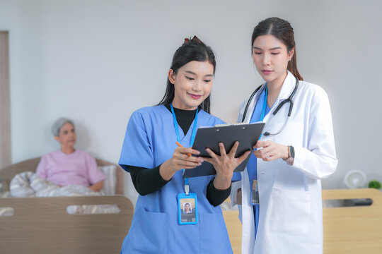 A female nurse in blue scrubs and a female doctor in a white coat share a conversation mid consultation with the tablet held between them in a modern elderly care home in Thailand.