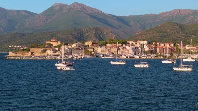 Saint Florent Corsica 4K drone video. Low altitude flight between sailboat masts to marina at sunset with yachts anchored near town surrounded by mountains. Scenic travel b roll stock footage