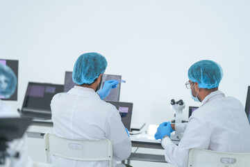 Lab technicians review results on a tablet by the microscope in a bright research laboratory while...