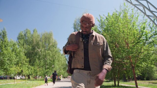 Close up of man walking outdoors in bright sunlight adjusting his jacket and carrying bag. Man wears casual clothing including cap with sunglasses hanging from neck interacting naturally