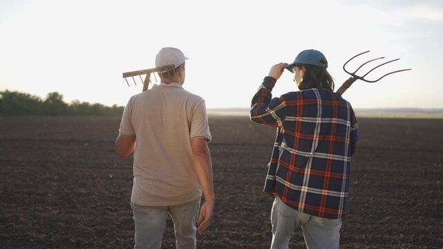 Farmer farm ground pitchforks. A man and a young man walk together across the field, discussing their farming plans. The man carries a rake while the young man holds a pitchfork.
