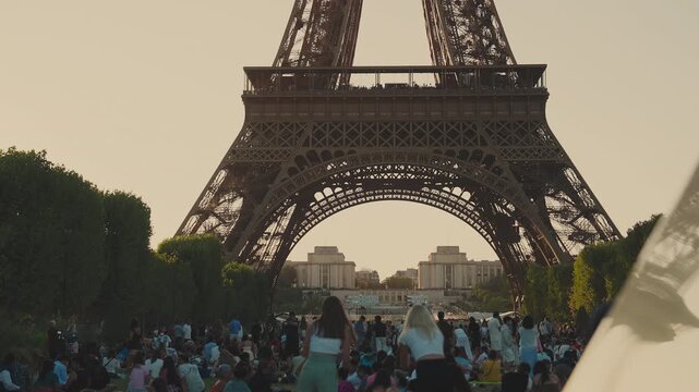 Static wide shot of people and tourists relaxing on the grass at Champ de Mars park with the Eiffel Tower base framed against a warm golden sunset sky in Paris, France. High quality 4K B-roll video.