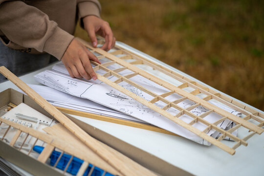 Teenager assembling wooden model airplane wing from balsa parts on table with blueprints. Hobby of aircraft modeling and engineering education for kids. Handmade construction and creativity concept.