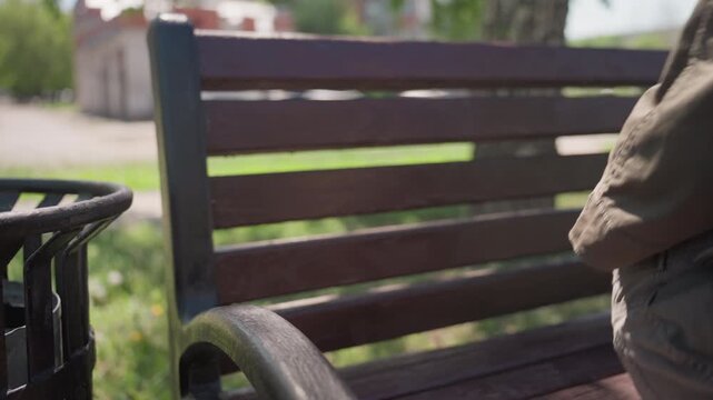 Close up of brown backpack with metal zippers and keyring attachments resting on outdoor bench next to black trash can. scene shows bag in focus while background elements like greenery
