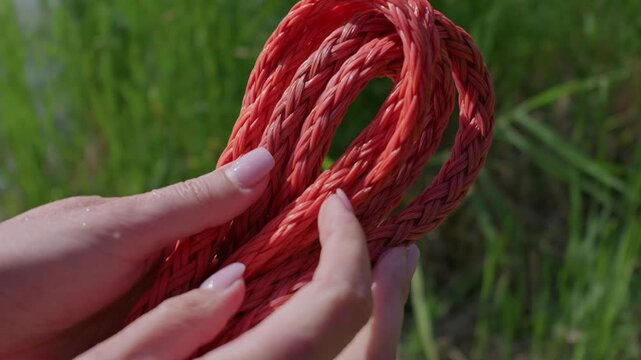 A woman hands hold coiled red braided synthetic rope against bright green grass outdoors. Concept of durable marine equipment, reliable utility gear, and braided cordage strength.