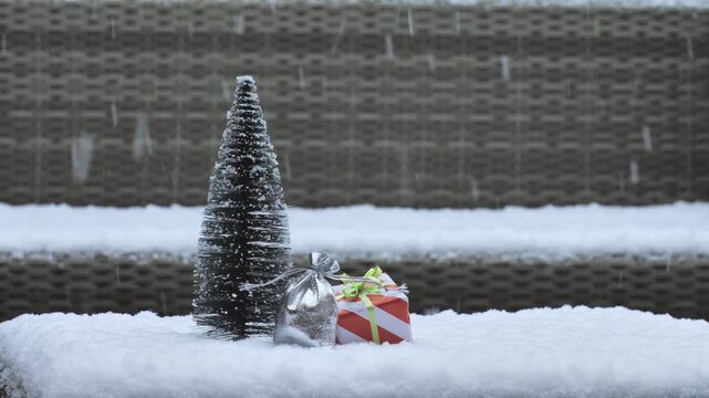 Snow gently falls around a small, snowy fir tree. Beside it sits a wrapped gift box with red
