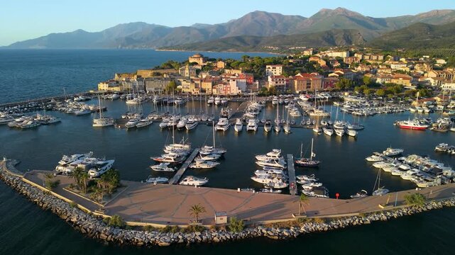 Saint-Florent Corsica drone descending flight between docked yachts marina towards historic town in golden hour light, luxury harbor central aisle. Cinematic 4K low-altitude drone footage