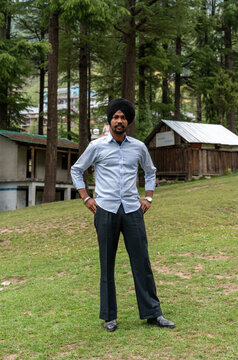 Full-length portrait of a young Sikh man wearing a black turban, light blue shirt, and black pant, standing with hands on his hips on green grass at Devidarh park with pine trees in the background.