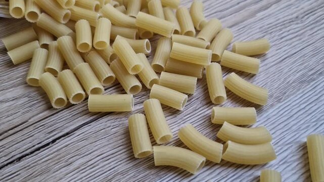 A close-up view of uncooked macaroni pasta scattered on a wooden surface. The pasta is short, tubular, and yellow in color.