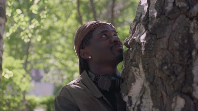 Close up of man wearing brown cap and jacket observing tree in park with green foliage soft lighting closeup greenery outdoor nature observation silence