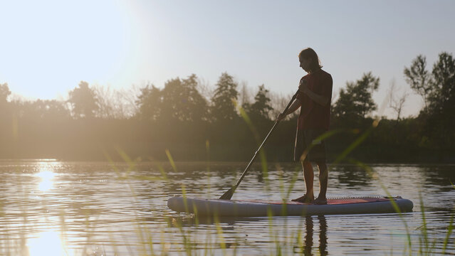 Supboard boad sun. A man paddles on a tranquil lake. The man enjoys paddleboarding amid serene water, while sunlight glistens on the lake's surface.