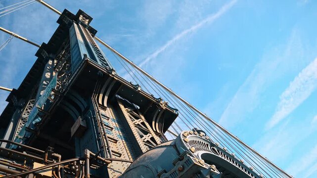 Manhattan Bridge steelwork beneath a blue sky. The low angle view frames a tower, suspension lines and open copy space.