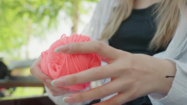 Close up of young woman untangling bright pink yarn in soft natural light. focus remains on her hands and texture of both skin and fabric with green foliage blurred in background. Soft