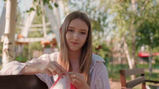 Close up of young woman crocheting fabric with red border in park setting holding crochet hook and yarn. Background features green trees playground equipment like swings or slide visible