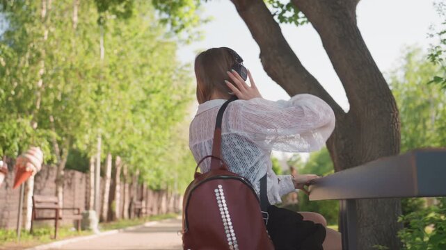 Close up of young woman wearing headphones and carrying red backpack transitioning from sitting at outdoor table to walking along tree lined path in park. Background features string lights