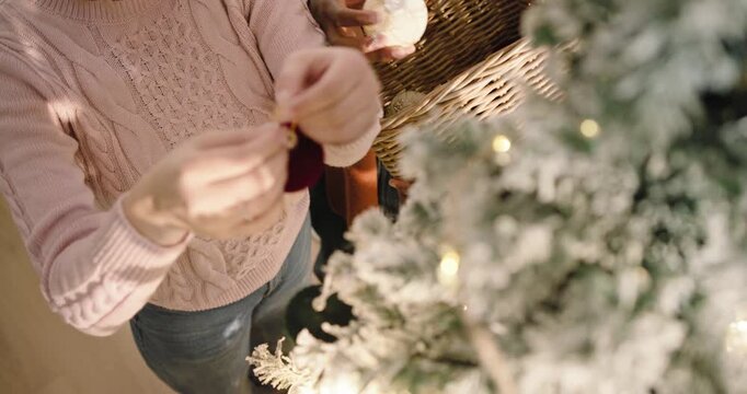 Reaching into wicker basket, two adults are hanging red velvet baubles on frosted tree for holidays