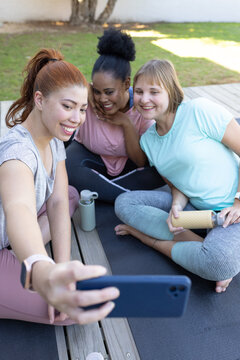 Diverse female friends sitting on deck mat leaning toward phone taking selfie with water bottles