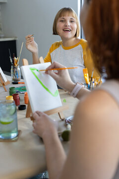 Two women sitting at art studio table painting on small canvases with easels, jars, brushes