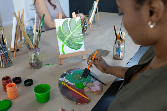 African American women painting at communal table in studio, holding foam brush over palette