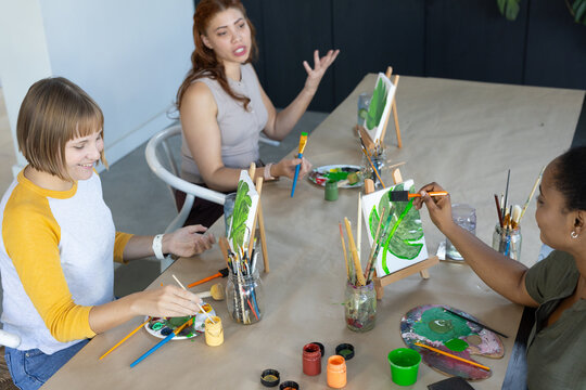 Three female artists painting together at studio table with easels, brushes and paint jars