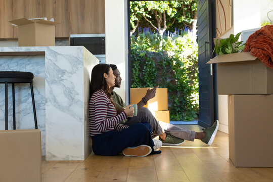 Diverse couple sitting on floor at home next to marble island holding ceramic mug unpacking boxes