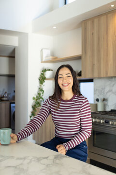 Woman sitting on kitchen island holding light-green mug smiling at camera in striped top and jeans