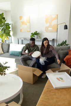 Diverse couple unpacking white plate, bubble wrap and cardboard boxes on gray sofa at home