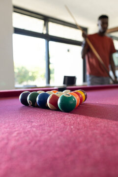 African man wearing rust T-shirt holding cue in game room near racked balls on burgundy felt