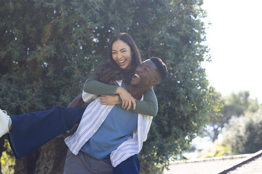 Diverse couple laughing while man carrying woman on back at park by evergreen tree, striped shirt