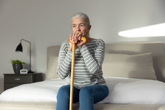 Senior female sitting on bed edge in bedroom holding wooden cane and gazing toward light