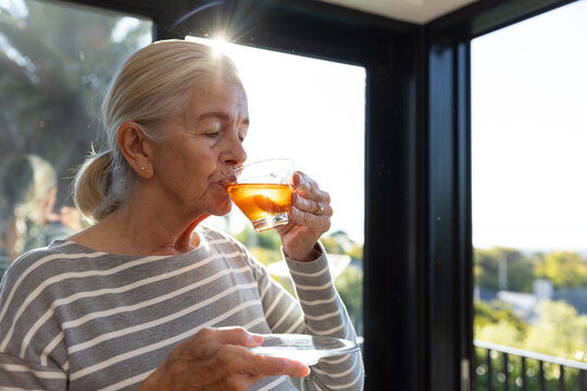 Senior woman wearing striped shirt holding glass teacup and saucer, sipping tea near glass doors