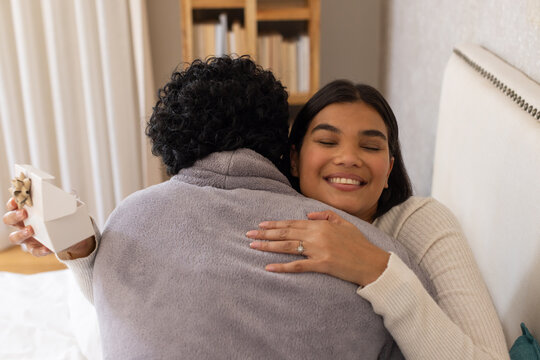 African American woman wearing ribbed sweater embracing partner on bed holding box with gold bow