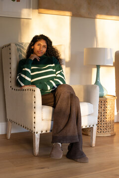 African American woman sitting in beige chair at home, wearing striped sweater, teal lamp visible