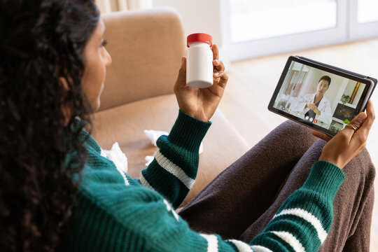Adult female sitting on beige sofa in green sweater holding pill bottle using tablet