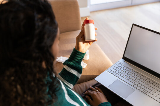 African American woman in green sweater holding and checking small white bottle by laptop at home