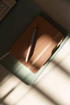 Tan notebook resting with white stylus on mint sleeve, blinds casting shadows across cream desk