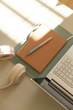 Tan leather notebook resting on green pad near window, pen lying diagonally, laptop and headphones