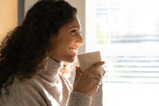 Adult African woman holding mug with rings, wearing knit sweater, looking toward window blinds